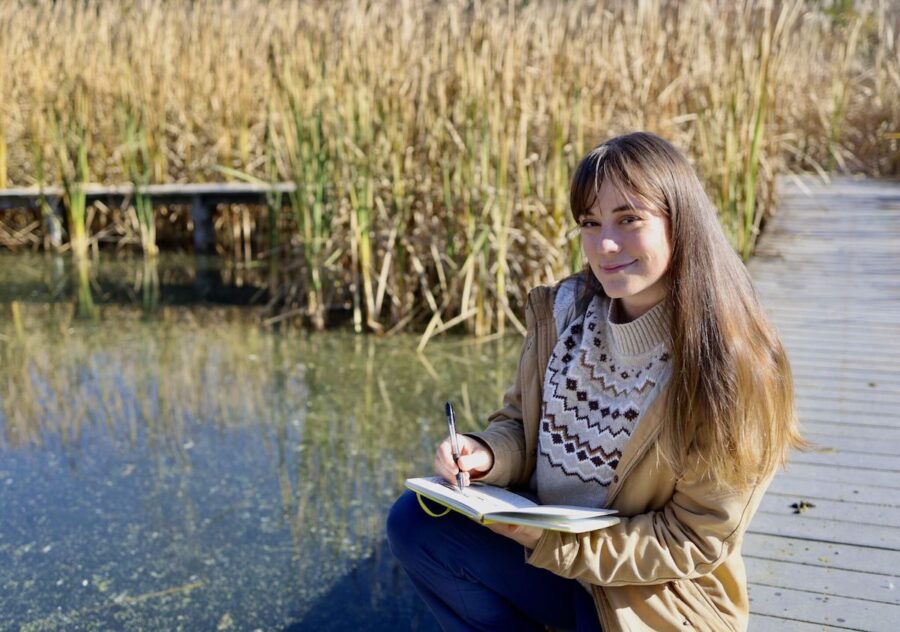 Photo of a woman on pier near a lake drawing in a journal