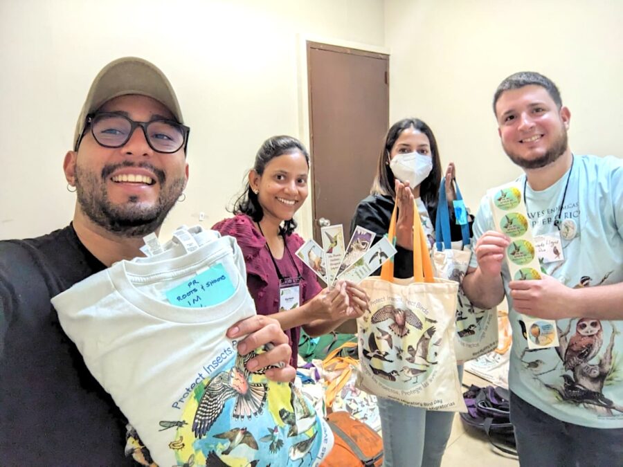 Selfie of group of four people holding t-shirts, tote bags, bookmarks, and stickers.