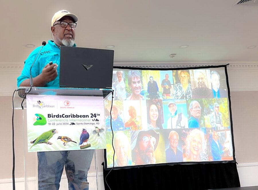 Man standing in front of an acrylic podium with laptop open. Behind him is a projector screen with pictures of his colleagues.